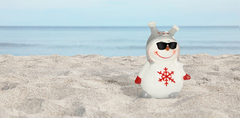 Cute small ceramic snowman with sunglasses on sandy beach near sea, space for text. Banner design