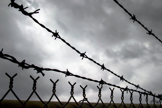 Horizontal close-up photo of an old and rusty chainlink fence topped with strands of barbed wire with dark storm clouds in the background.