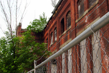 Horizontal photo of a cyclone fence in front of an old abandoned and boarded up red brick factory.