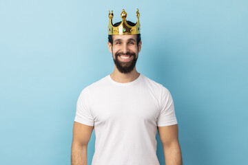 Portrait of man wearing white T-shirt and crown on head, smiling, concept of self confidence in success, self-motivation and dreams to be best. Indoor studio shot isolated on blue background. - obrazy, fototapety, plakaty