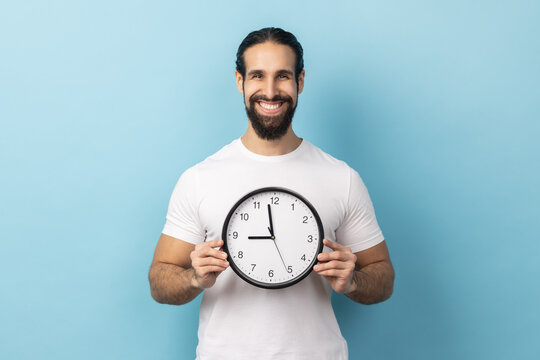 Portrait of positive optimistic man with beard wearing white T-shirt holding big wall clock, looking at camera with pleasant smile, time to go. Indoor studio shot isolated on blue background.