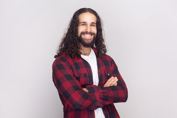 Portrait of satisfied cheerful handsome bearded man with long curly hair in checkered red shirt looks at camera, keeps hands folded. Indoor studio shot isolated on gray background.