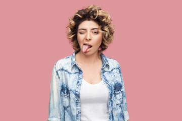 Portrait of funny childish attractive woman with curly hairstyle wearing blue shirt standing looking at camera showing tongue out. Indoor studio shot isolated on pink background. - obrazy, fototapety, plakaty