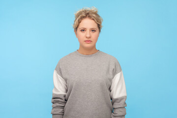 Portrait of attractive serious strict blonde woman in sweatshirt looking at camera with bossy expression, standing with confident look. Indoor studio shot isolated on blue background.