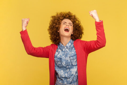 Portrait of woman with Afro hairstyle clenches fists with positive expression, rejoices wonderful news, smiles happily, squints face with pleasure. Indoor studio shot isolated on yellow background.