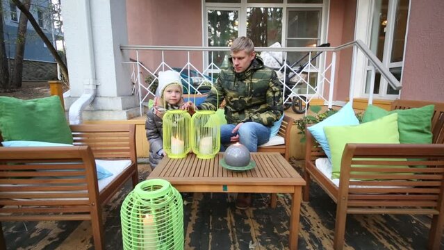 Father and daughter are sitting on terrace near house, lamps with candles