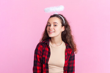 Portrait of smiling angelic teenage girl with wavy hair in red checkered shirt and nimb over head looking smiling at camera. Indoor studio shot isolated on pink background.