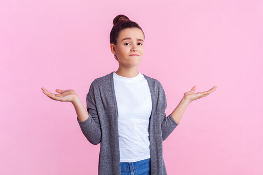 Portrait Of Confused Teenage Girl With Bun Hairstyle In Casual Clothes Standing With Shrugging Shoulders Having Uncertain Facial Expression. Indoor Studio Shot Isolated On Pink Background.