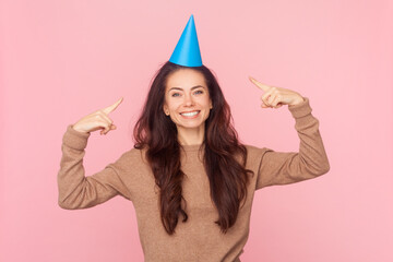 Portrait of smiling festive birthday woman pointing at her blue party cone, celebrating, expressing...