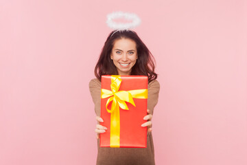 Portrait of joyful happy woman with wavy hair and nimb over head giving red wrapped present box, congratulating, wearing wearing brown pullover. Indoor studio shot isolated on pink background