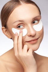 Under eye masks for puffiness, wrinkles, dark circles. Closeup portrait of beautiful woman applying gold eye patches. Indoor studio shot isolated over gray background.