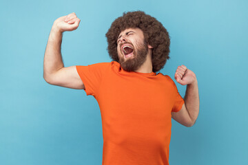 Portrait of sleepless man with Afro hairstyle wearing orange T-shirt yawning and raising hands up, feeling fatigued of overtime work. Indoor studio shot isolated on blue background. - obrazy, fototapety, plakaty
