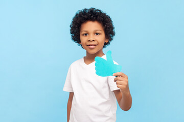 Portrait of cheerful positive little boy with curly hair showing paper thumb up internet trends...