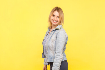 Side view portrait of smiling satisfied blonde woman wearing denim jacket looking at camera with toothy smile. Indoor studio shot isolated on yellow background.