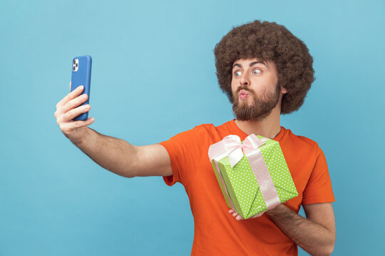 Portrait of romantic man with Afro hairstyle wearing orange T-shirt having video call with gift box, boasting his gift to followers, sending air kisses. Indoor studio shot isolated on blue background.