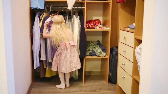 Little girl in a pink dress sorts out clothes on hangers in wardrobe