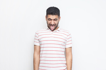 Portrait of foolish crazy childish bearded man wearing striped t-shirt standing with foolish facial expression, sticking tongue out. Indoor studio shot isolated on gray background.