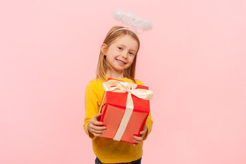 Portrait of joyful friendly blonde little girl with nimb over head holding red wrapped gift box, giving present, congratulating, wearing yellow jumper. Indoor studio shot isolated on pink background.