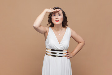 Woman holding hand above eyes and peering into distance, looking far away, expecting and searching someone on horizon, wearing white dress. Indoor studio shot isolated on light brown background.