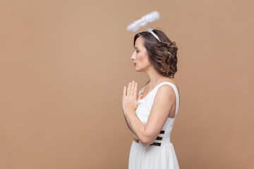 Portrait of hopeful middle aged woman with wavy hair and nimb over head, standing with palms together, praying gesture, wearing white dress. Indoor studio shot isolated on light brown background.