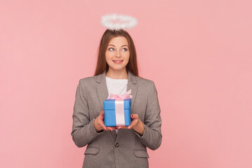 Portrait of dreaming smiling woman with brown hair with nimb over head holding gift box, making wish on birthday, wearing business suit. Indoor studio shot isolated on pink background.