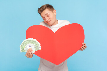 Portrait of rich delighted man wearing white t-shirt holding big red heart and fan of euro banknotes, love to money. Indoor studio shot isolated on blue background