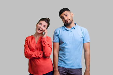 Portrait of couple man and woman cheating using phone and smiling happily, jealous guy waiting his wife talking cell phone. Indoor studio shot isolated on gray background.