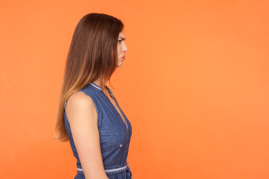 Side view portrait of sad unhappy brunette woman wearing denim dress looking ahead being in bad mood offended stressed annoyed expression. Indoor studio shot isolated on orange background.
