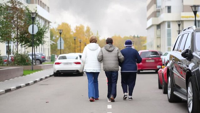 Three senior people walk on road along parked cars in autumn day