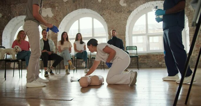 Close-up shooting: a bearded man brunette in a gray T-shirt starts a practical lesson showing first aid together with a professional doctor a woman in a white uniform with a mannequin in front of the