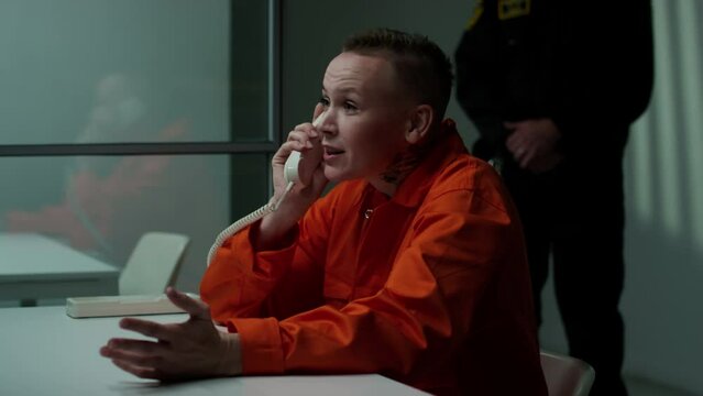 Nervous female prisoner in orange jail uniform sitting in meeting room and having emotional talk with visitor via telephone