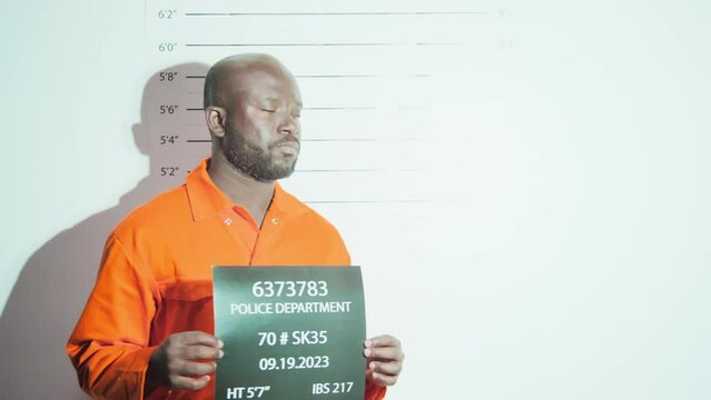 African American suspect in orange jail uniform standing against height chart and holding sign with number while photographer taking mugshots