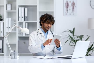 Young medical practitioner concentrated on digital tablet while working in a modern clinic. Represents healthcare technology. - obrazy, fototapety, plakaty