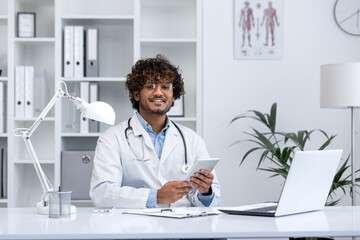 Cheerful young male healthcare professional with stethoscope sitting at his desk in a medical...
