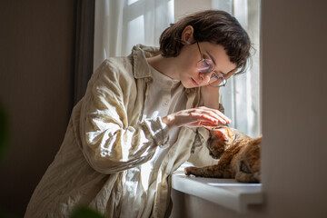 Teen girl feeling lonely, stressed in need of friendship, emotional support, tactile contact, standing near windowsill, caressing, stroking cat devon rex, thinking of life. Pet in role of antistress 