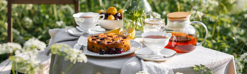 Elegant glamour table setting outdoor in the garden. White porcelain cups, teapot with herbal tea, homemade cherry sweet pie. Family tradition, beautiful place outside. Flower meadow, candles. Banner