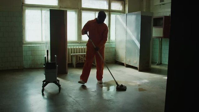Full shot of African American male prisoner in orange uniform mopping floor while doing cleaning chores