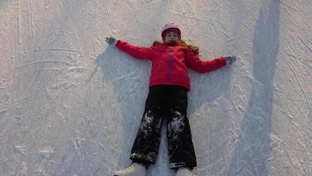 Girl in helmet, coat and skates lie on skate rink and wave hand and legs