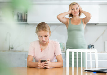 Unconcerned teen girl fixated on phone sitting at table in home kitchen while dissatisfied mother...