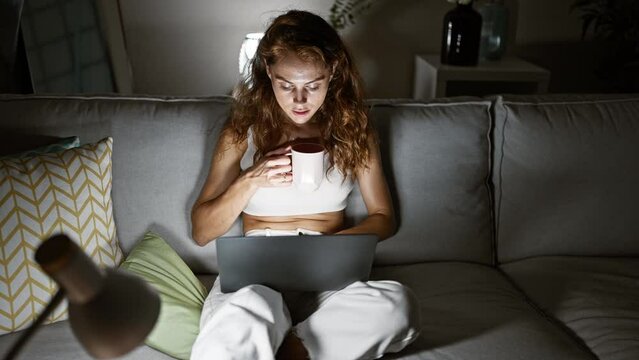 A young woman works on her laptop at night in the living room, enjoying a cup of coffee on a comfortable couch, reflecting a cozy home environment.