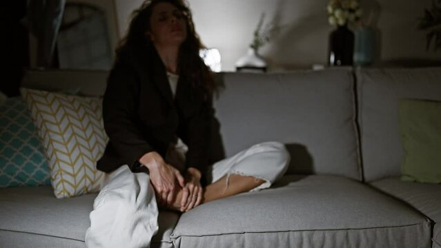 Tired young woman sitting on couch at home, relaxing in a dimly lit cozy living room
