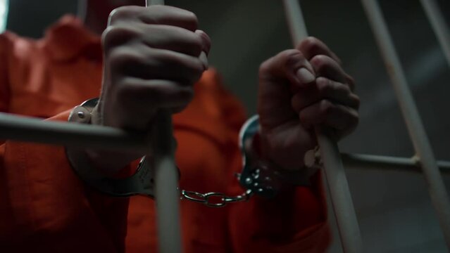 Close up low angle view of hands of female prisoner in handcuffs and orange jail uniform holding cell barsClose up low angle view of hands of female prisoner in handcuffs and orange jail uniform holdi