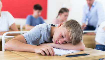 Fototapeta na wymiar Tired teen student sleeping at desk in classroom during lesson on blurred background of classmates ..