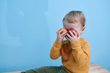 2 year old boy sitting holding red apples and closing eyes background blue wall