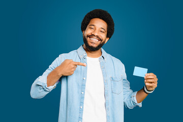 The African-American man points to a credit card with a confident smile standing isolated on blue, suggesting approval of a product or service, or recommending a financial tool or app.