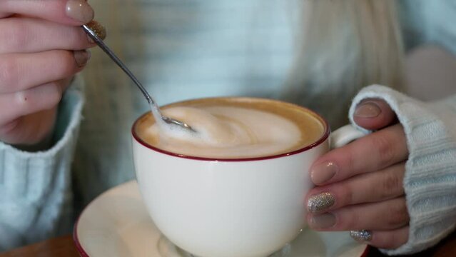 Young woman mixes cappuccino by spoon in hands with vanish on nails