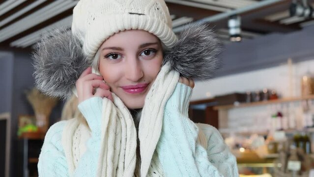 Blonde in cap holds scarf with fur near face and smiles in cafe