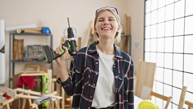 A smiling woman holding a drill in a bright carpentry workshop, portraying skill and confidence.