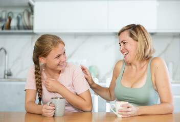 Smiling adult woman and teenage girl spending time together, engrossed in lively friendly conversation over cup of coffee at home kitchen table..
