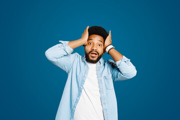 Young man in a denim shirt looks surprised with hands on his head against blue background. His...
